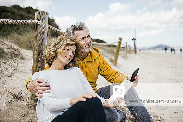 Lächelnder reifer Mann mit Frau  die am Strand ein Buch liest
