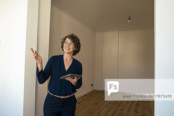Smiling real estate agent with tablet PC standing at doorway gesturing in new home