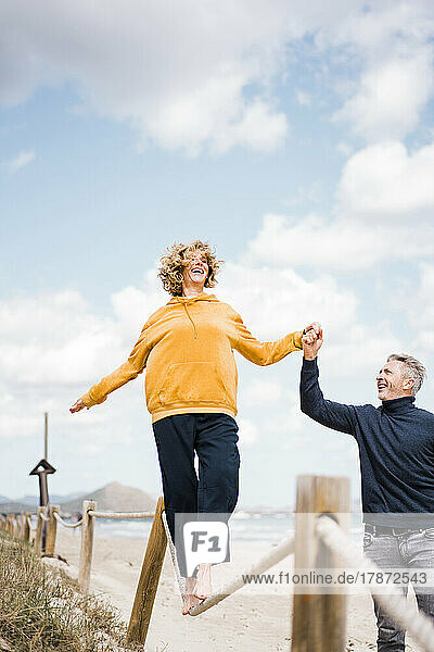 Glückliche Frau hält Händchen mit Mann  der am Strand auf einem Seil läuft
