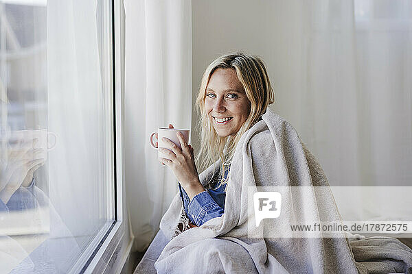 Young woman wrapped in blanket sitting near window holding cup at home