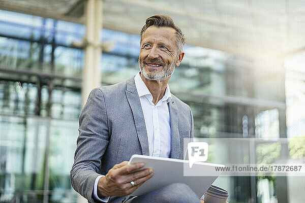 Smiling mature businessman with digital tablet in front of building