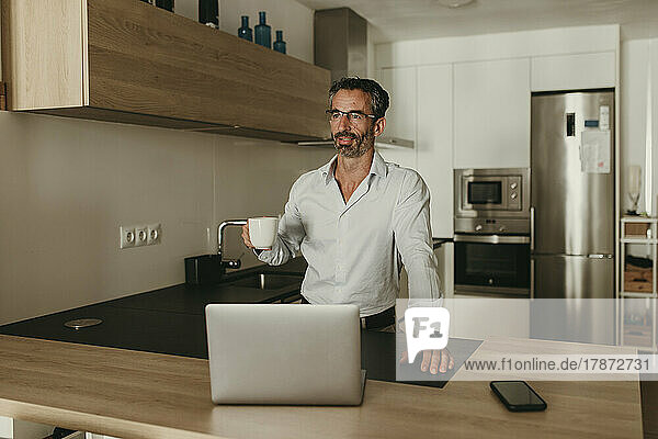 Businessman with coffee cup by smart phone and laptop at kitchen counter