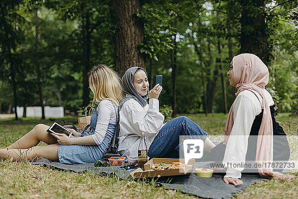 Young woman photographing friend sitting back to back at park