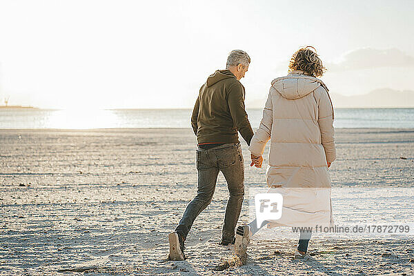 Älteres Paar hält sich an den Händen und geht an einem sonnigen Tag am Strand spazieren