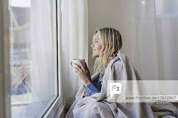 Smiling woman wrapped in blanket holding cup looking out of window at home