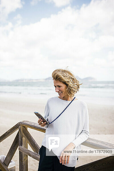 Smiling woman using smart phone leaning on railing at beach