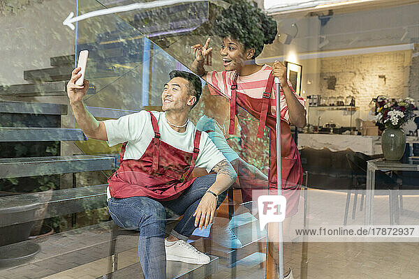 Lächelnder Mann macht Selfie mit dem Handy  während sein Kollege durch Glas gesehen wird