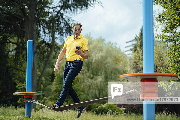 Surprised man looking at smart phone balancing on tightrope in park