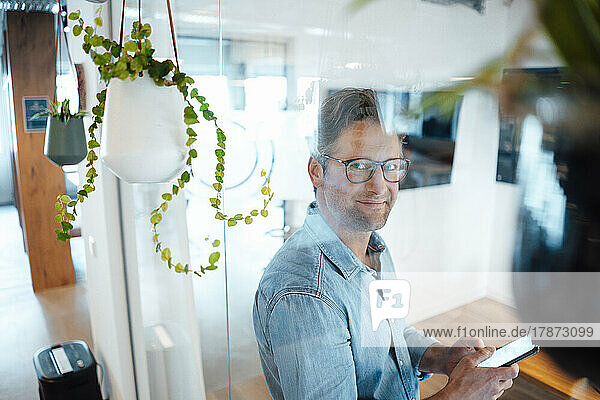 Smiling businessman with smart phone seen through glass wall in office