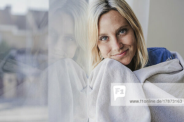 Smiling woman wrapped in blanket leaning on window glass