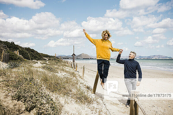 Happy mature woman walking on rope with man holding hand at beach