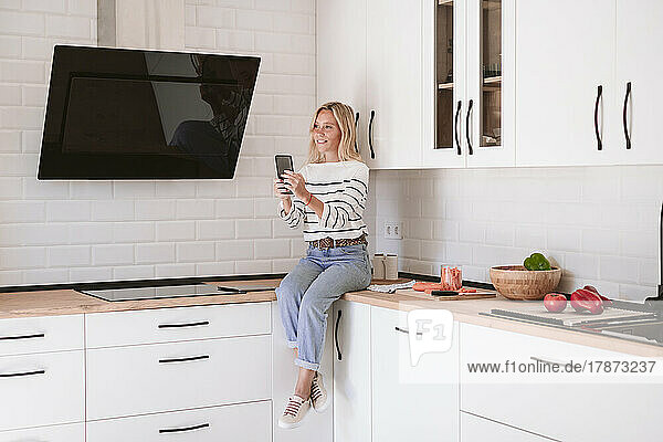 Smiling woman holding mobile phone on kitchen counter at home