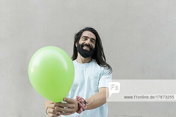 Glücklicher Hipster-Mann mit langen schwarzen Haaren  der vor der Wand einen grünen Ballon schenkt