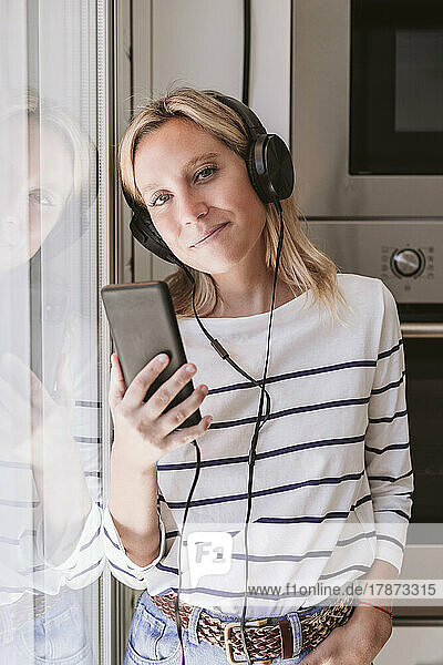 Smiling woman with mobile phone listening music in domestic kitchen