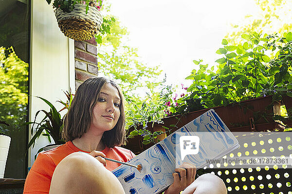 Teenagermädchen zeigt Aquarellmalerei auf Skizzenblock und sitzt auf dem Balkon