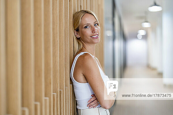 Smiling businesswoman with arms crossed standing by wall at workplace