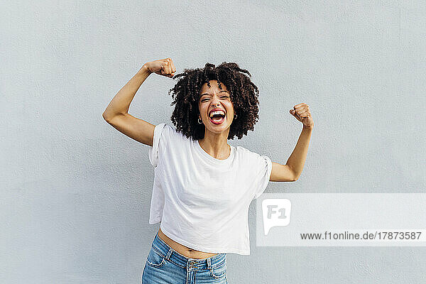Cheerful young woman flexing muscles in front of white wall