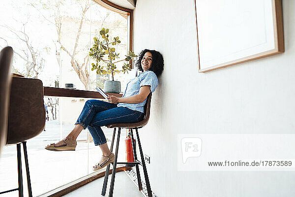 Happy woman with tablet PC sitting on chair by window at cafe