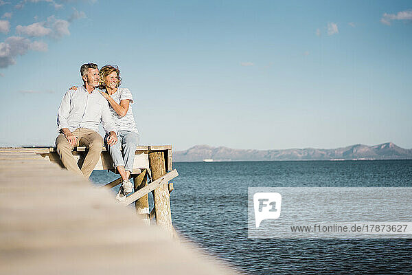 Glückliches älteres Paar sitzt auf einem Steg über dem Meer vor dem Himmel