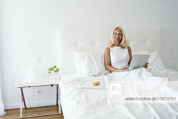 Happy woman with laptop sitting by breakfast tray on bed
