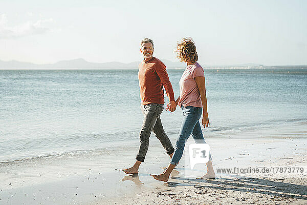 Happy mature couple holding hands walking on shore at beach