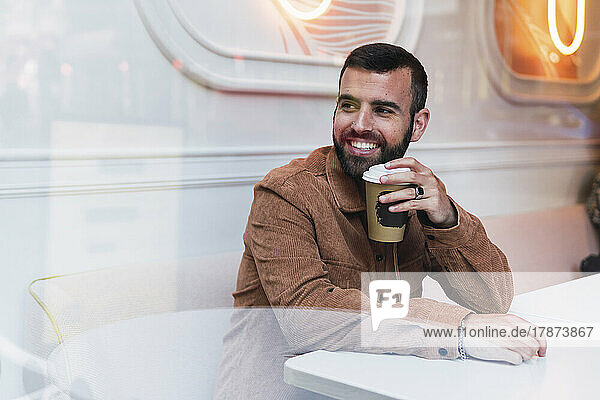 Smiling young man with disposable coffee cup sitting at table in cafe