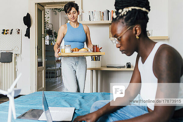 Young woman carrying breakfast on tray for girlfriend working at home