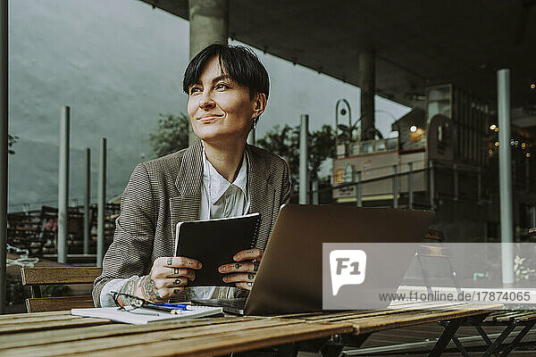 Smiling businesswoman with laptop and diary sitting at sidewalk cafe