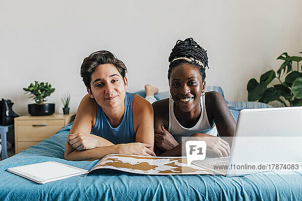 Lächelndes lesbisches Paar mit Laptop und Tablet-PC auf dem Bett liegend
