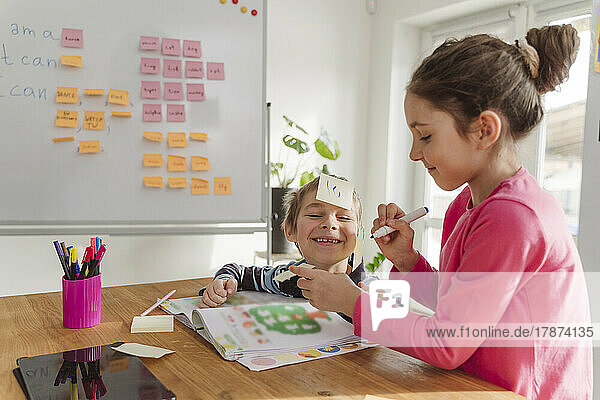 Smiling boy with adhesive note on forehead by sister studying at home