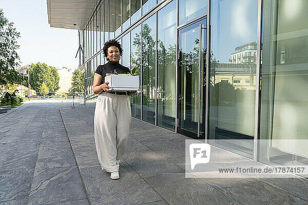 Smiling businesswoman holding box walking on footpath