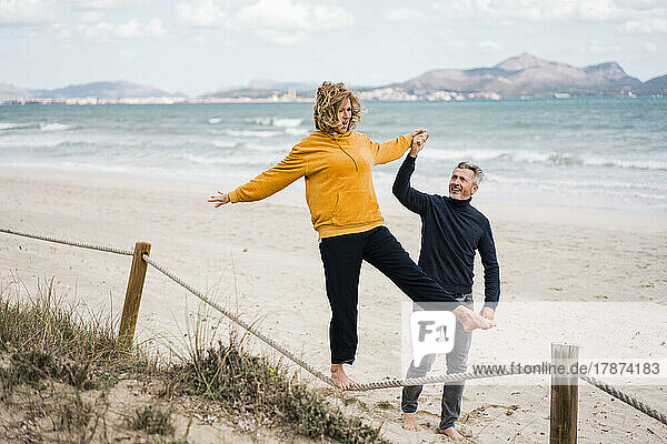 Smiling man holding hand of woman walking on rope at beach