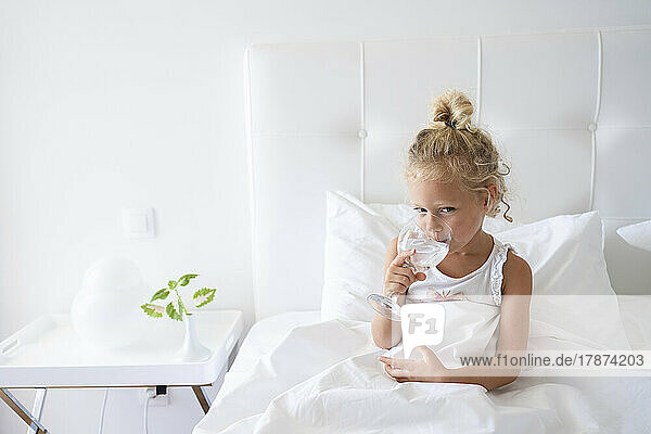 Blond girl drinking water sitting in bed at bedroom