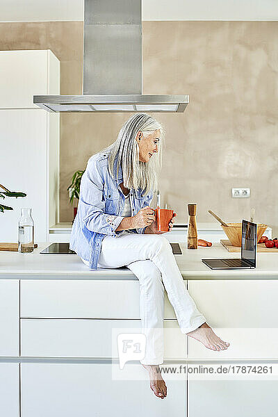 Frau mit Tasse und Laptop sitzt auf der Küchentheke