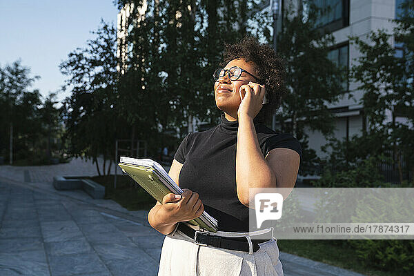 Smiling young businesswoman with eyes closed holding laptop talking on mobile phone