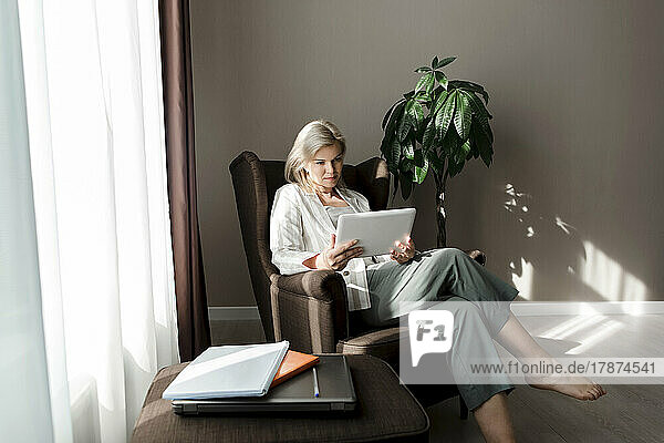 Reife Frau sitzt zu Hause im Sessel und benutzt einen Tablet-PC