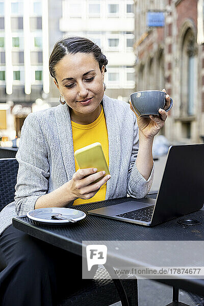Lächelnde Geschäftsfrau hält Kaffeetasse mit Smartphone im Straßencafé in der Hand