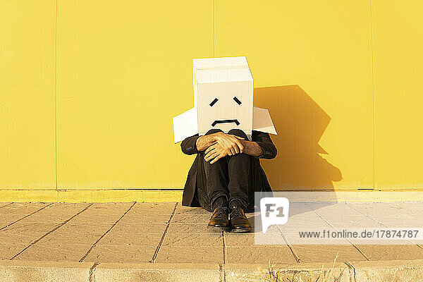 Man wearing box with sad face sitting in front of yellow wall on footpath