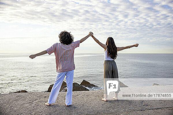 Couple holding hands standing with arms outstretched on pier looking at sea