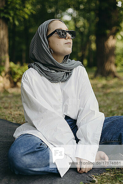 Young woman wearing sunglasses and hijab sitting cross-legged in park
