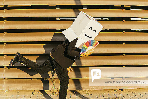 Playful businessman wearing box with face holding rainbow color hand fan on sunny day