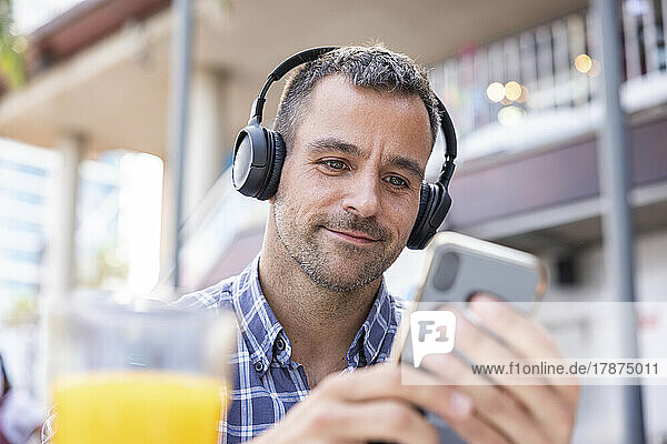 Smiling mature man using smart phone listening music through headphones at cafe