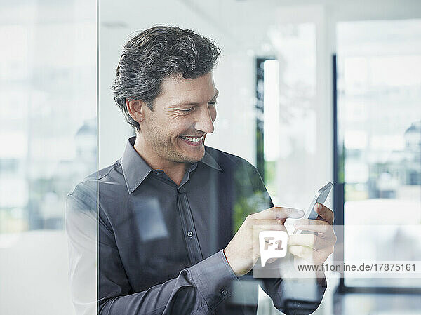 Happy businessman using smart phone seen through glass
