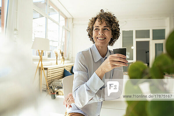 Happy businesswoman with disposable coffee cup sitting at desk in office