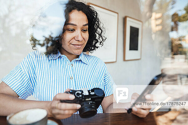 Smiling woman with camera using smart phone at table in cafe
