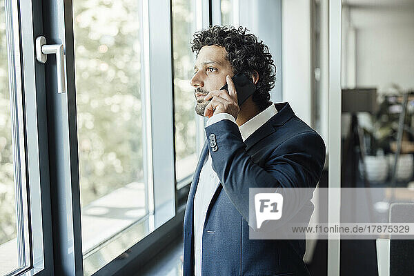 Businessman talking on smart phone looking out through window in office