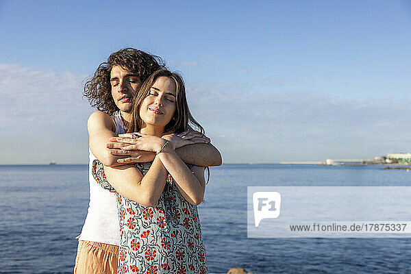 Young couple standing with arms around in front of sea on sunny day