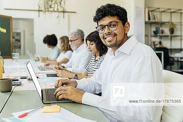Smiling businessman with laptop sitting by colleagues at office