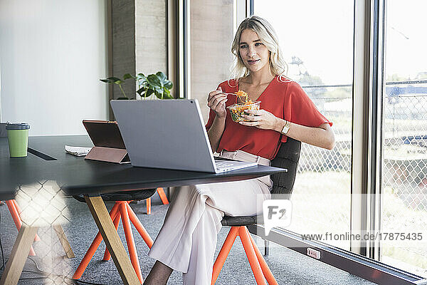 Businesswoman having salad looking at laptop sitting on chair in office