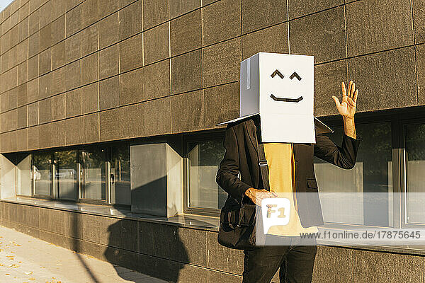 Businessman wearing box with smiley face waving hand at footpath on sunny day